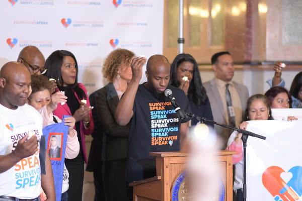 A Black man stands a podium, head cast down with his eyes closed. He is wearing a t-shirt that reads “Survivors Speak”. In his right hand he holds a candle as a group of people behind him, also holding candles, look solemn.