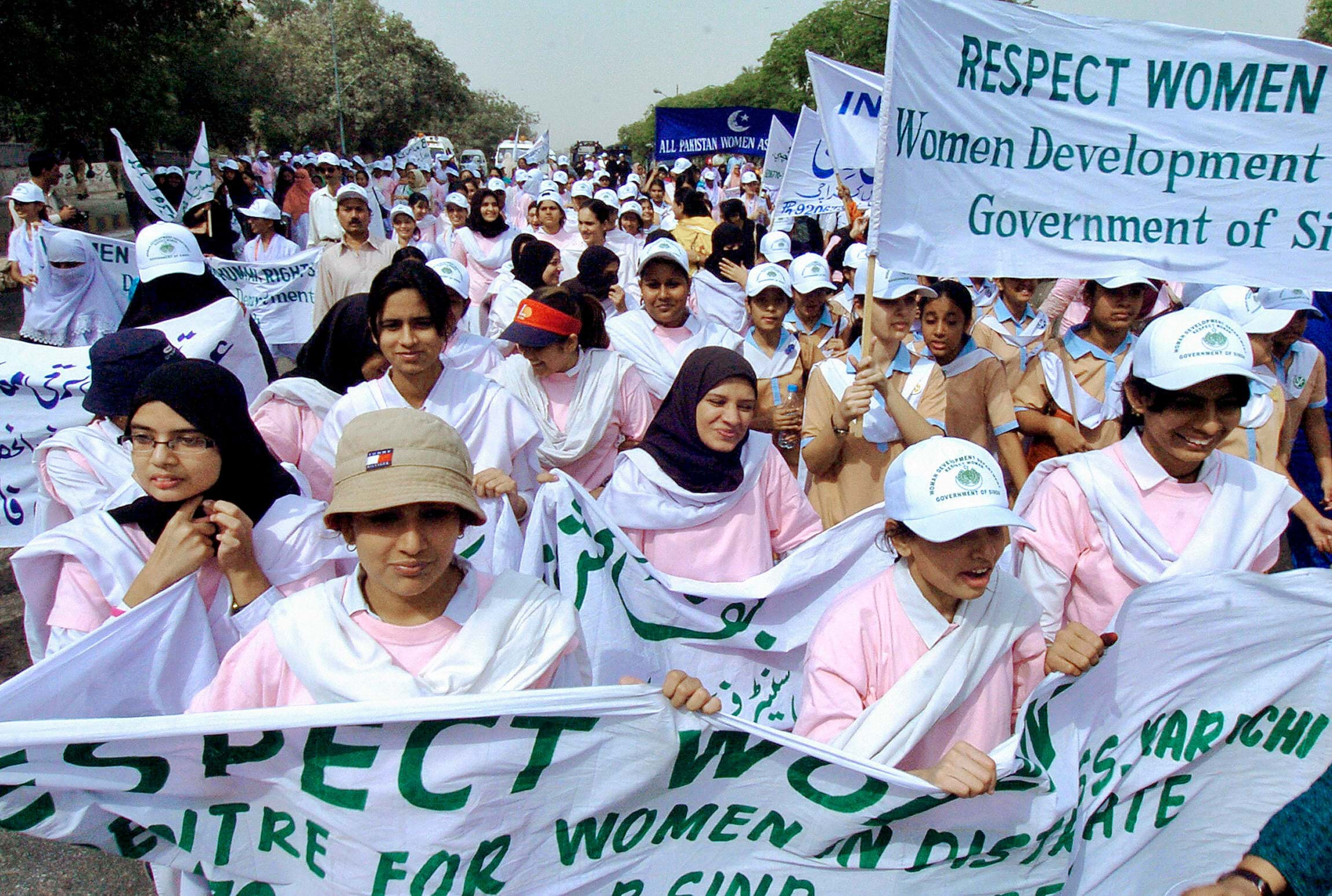 Protesters wearing pink polo shirts and a mixture of hats, hijabs, and no head covering march with protest signs.