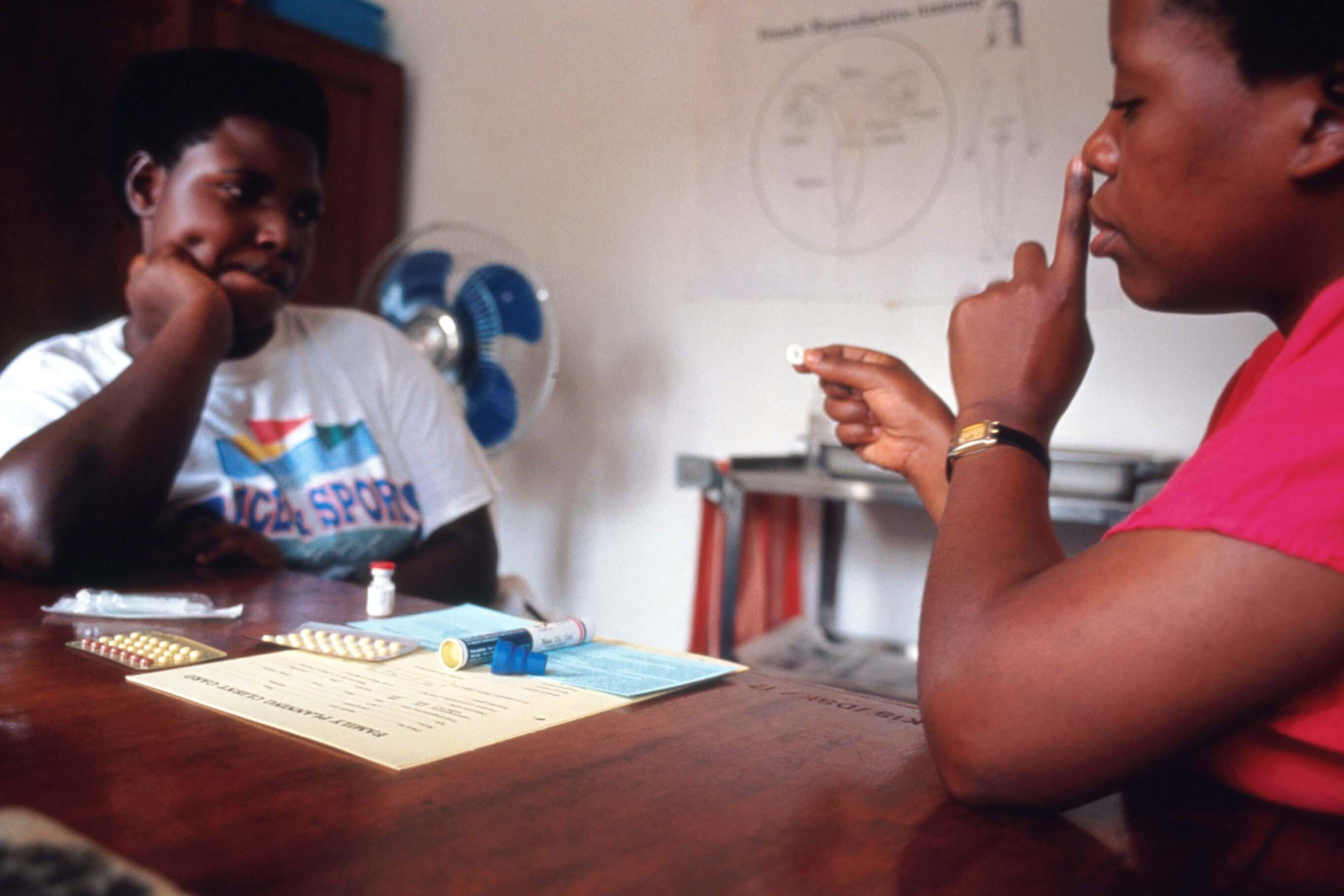 Two black people sit across from each other at a table. A poster and medication are in front of them.