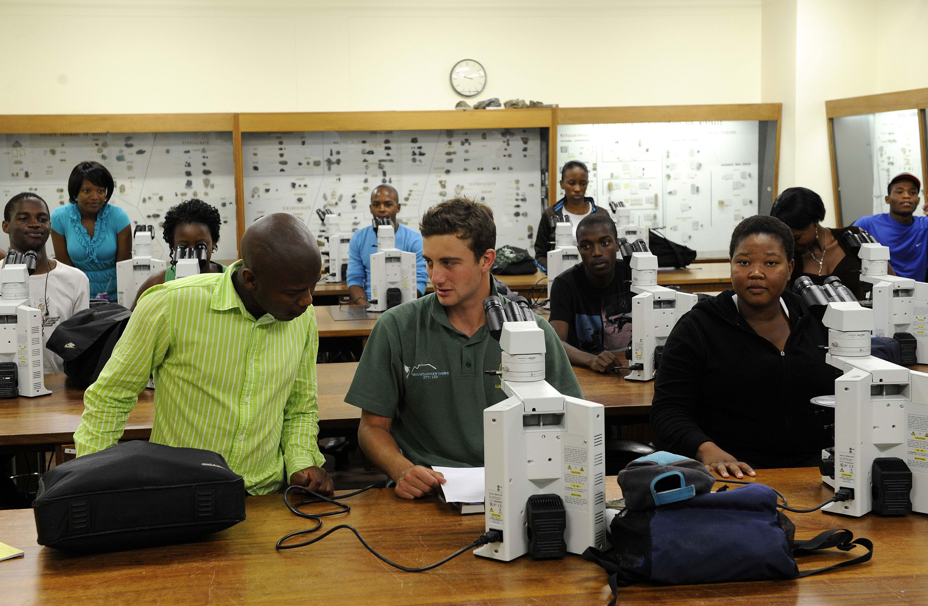 A mix of Black men and Black women sit at long tables in a classroom. A Black person in a neon green button-down shirt confers with a white man in a dark green polo.