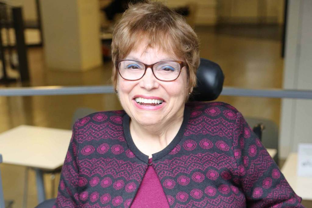 Judy Heumann has short, auburn hair and wears dark, tortoise shell glasses and a black and fuchsia patterned cardigan. She smiles while seated in a powered wheelchair.