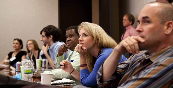 A row of people sit at a table focused pensively on something at the front of the room.