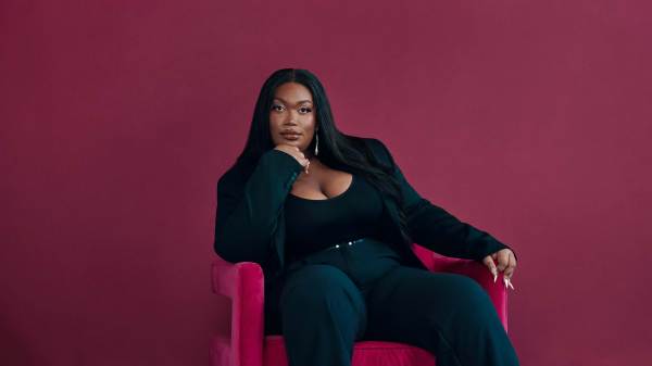 Before a vibrant magenta backdrop, Jordyn Jay, a Black trans femme, leans elegantly on a plush, pink chair, her thoughtful pose accented by a hand delicately supporting her chin. Draped in a sleek, black pants suit, her long hair cascades from a central part, providing contrast to the surrounding hues.