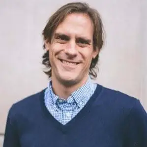 Matt Hansen wearing a blue & white stripped button down shirt with a blue v-neck sweater over it against a light background.