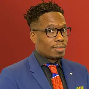 Matt Mitchell wearing a black shirt, orange and blue tie and blue blazer against a maroon background,