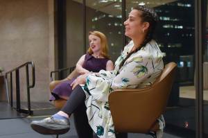 Rebecca Cokely, a little person, sitting in a brown leather chair in conversation with Maysoon Zaiyd, an average-size person against the window of the city skyline. They are both smiling.