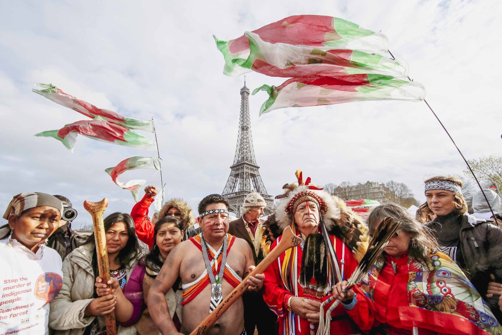 Indigenous Peoples from around the world wearing traditional clothes on a ferry on the Paris Seine River, behind them is the Eiffel Tower. With their canoe destroyed during COP21 in Paris, the alliance boarded a ferry on the Seine and invited Indigenous leaders from the North Pacific and the media to join their #PaddletoParis action. This ferry ride became the iconic image published on news outlets worldwide when Indigenous rights were recognized in the Paris Climate Change Agreement.